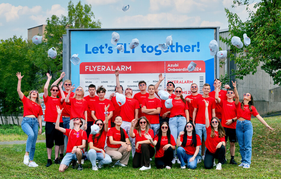 Gruppe junger Menschen in roten T-Shirts wirft weiße Kappen in die Luft, im Hintergrund ein Schild mit der Aufschrift Let’s FLY together! und Azubi Onboarding 2024.
